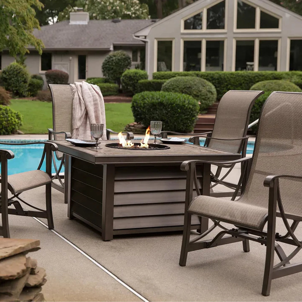 Outdoor dining setup by the pool with a square fire pit table and four high-back chairs, set against a backdrop of a manicured garden and a large house with a sunroom, creating a luxurious and inviting ambiance