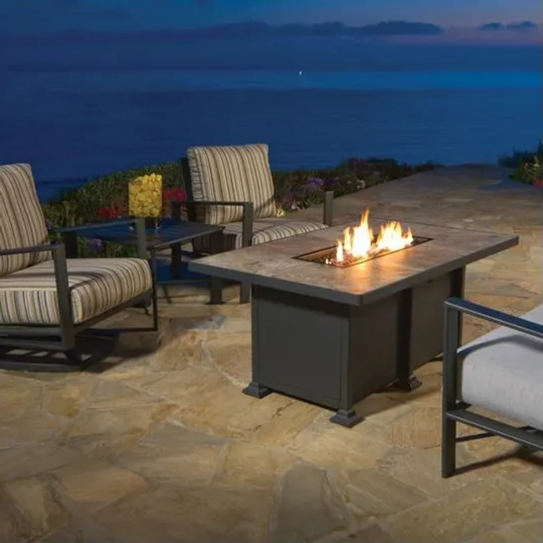 Modern rectangular fire pit table with a stone top and linear flame, set on a stone patio overlooking the ocean at dusk, surrounded by striped cushioned outdoor chairs