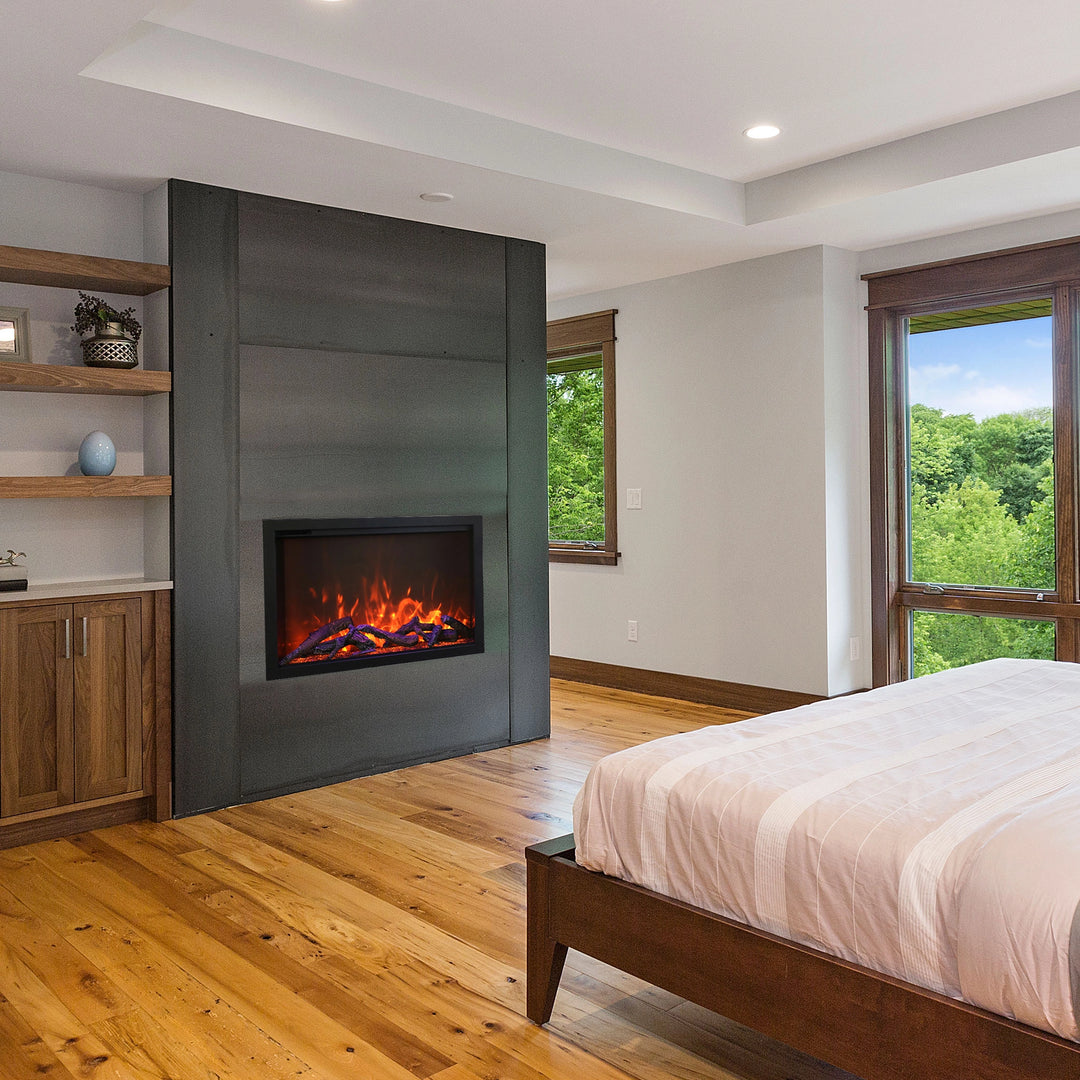 The Amantii Traditional Bespoke Smart Electric Fireplace installed in a modern bedroom with wooden flooring and large windows. Its realistic flame effects and glowing embers create a warm and inviting atmosphere, enhancing the room's natural aesthetic.