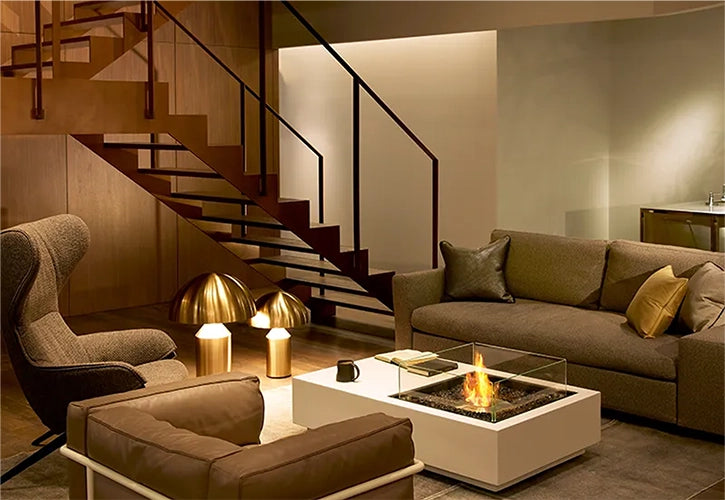 Modern living room featuring a fire pit integrated into a white coffee table, surrounded by neutral-toned furniture, warm lighting, and a sleek staircase, creating a cozy and sophisticated atmosphere