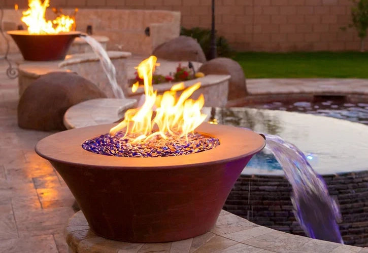 Backyard setup with copper fire bowls filled with blue fire glass and integrated water features, placed around a pool area with decorative stonework