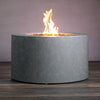 Beton 42-inch Graphite Round Concrete Gas Fire Pit with Access Door, displayed with flames on for a cozy ambiance. Its smooth graphite-colored finish complements any outdoor space. The round cylindrical shape offers a clean, modern look, making it perfect for backyard entertainment areas. Includes an access door for propane tank storage