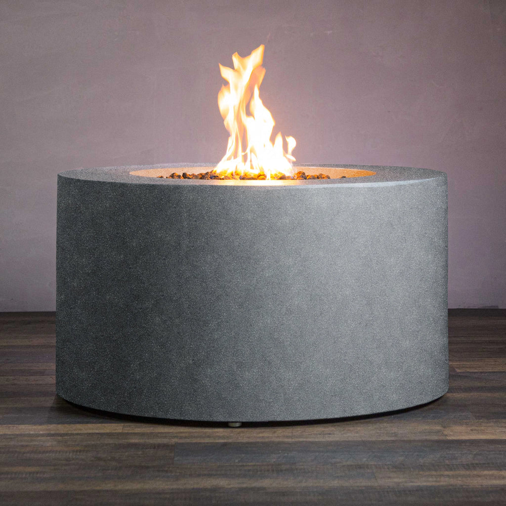 Beton 42-inch Graphite Round Concrete Gas Fire Pit with Access Door, displayed with flames on for a cozy ambiance. Its smooth graphite-colored finish complements any outdoor space. The round cylindrical shape offers a clean, modern look, making it perfect for backyard entertainment areas. Includes an access door for propane tank storage