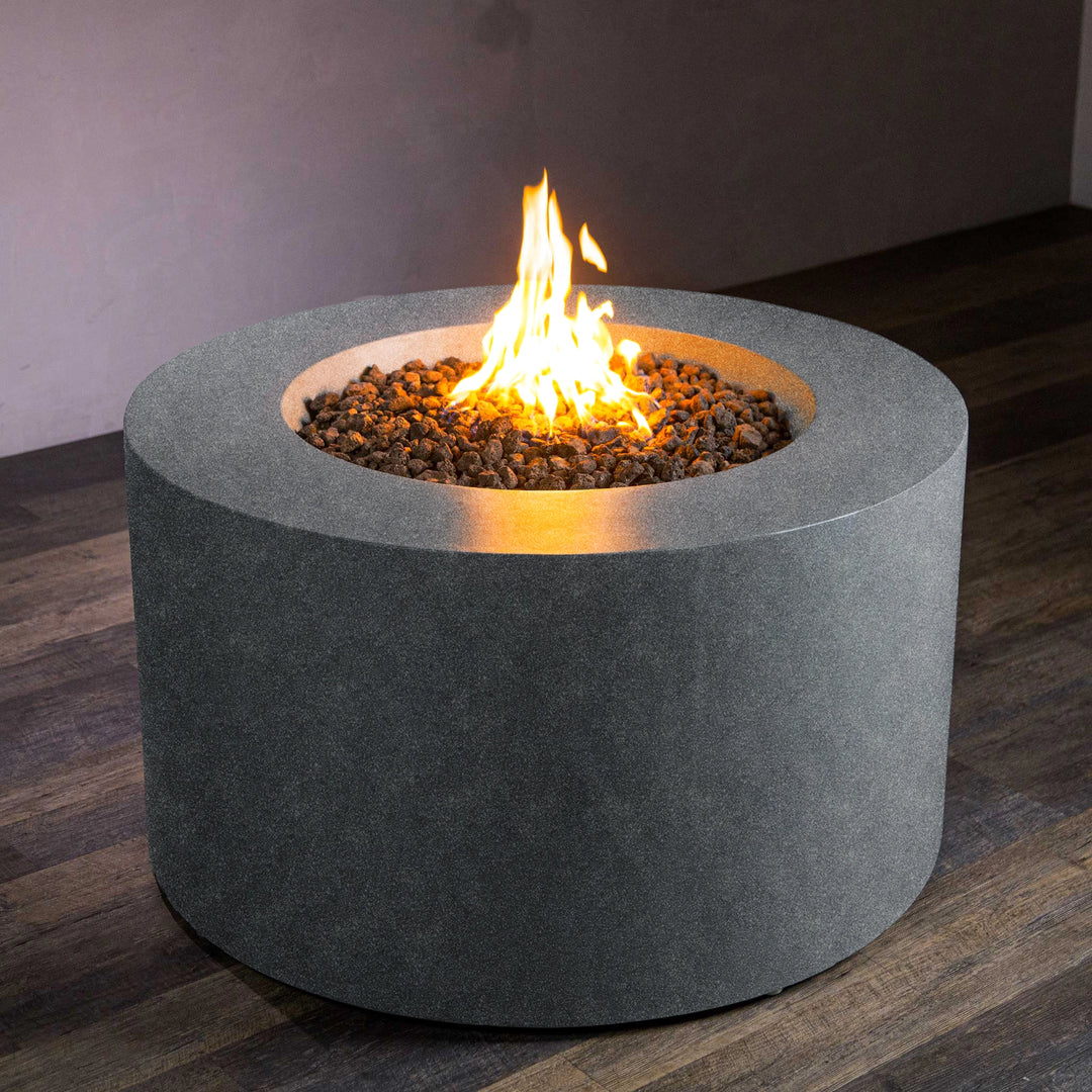 Beton 42-inch Graphite Round Concrete Gas Fire Pit with Access Door, featuring a modern cylindrical design and a strong graphite finish, shown with a bright natural gas flame and filled with lava rocks. Ideal for outdoor patio areas, it provides both warmth and a contemporary aesthetic. The access door allows for easy storage of a propane tank or maintenance access