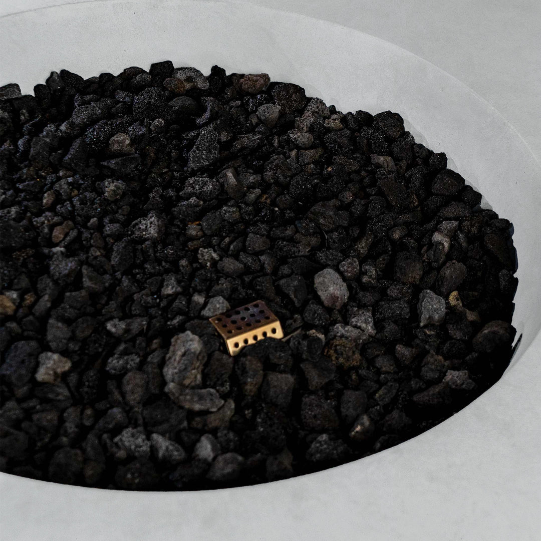 A close-up of the lava rocks used in the Beton 42" White Round Concrete Natural Gas Fire Pit. The durable black lava rocks are shown in the fire pit’s circular basin, designed to withstand high heat and enhance the aesthetic of the flame. This fire pit uses natural gas and is operated with a push-button ignition. The modern white concrete structure contrasts with the rugged texture of the lava rocks.