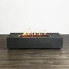 This studio view displays a modern fire pit with a sleek, graphite-colored finish resting on a dark wood floor. Bright orange flames rise continuously from a bed of black lava rocks, demonstrating the clean burn of a gas fire pit. The minimalist design of the linear fire pit is emphasized against a plain white background, showcasing the consistent flame pattern and architectural form without environmental distractions.