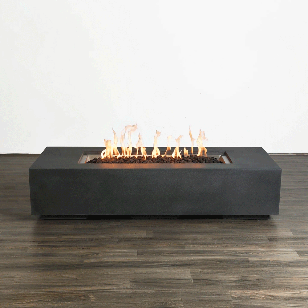 This studio view displays a modern fire pit with a sleek, graphite-colored finish resting on a dark wood floor. Bright orange flames rise continuously from a bed of black lava rocks, demonstrating the clean burn of a gas fire pit. The minimalist design of the linear fire pit is emphasized against a plain white background, showcasing the consistent flame pattern and architectural form without environmental distractions.