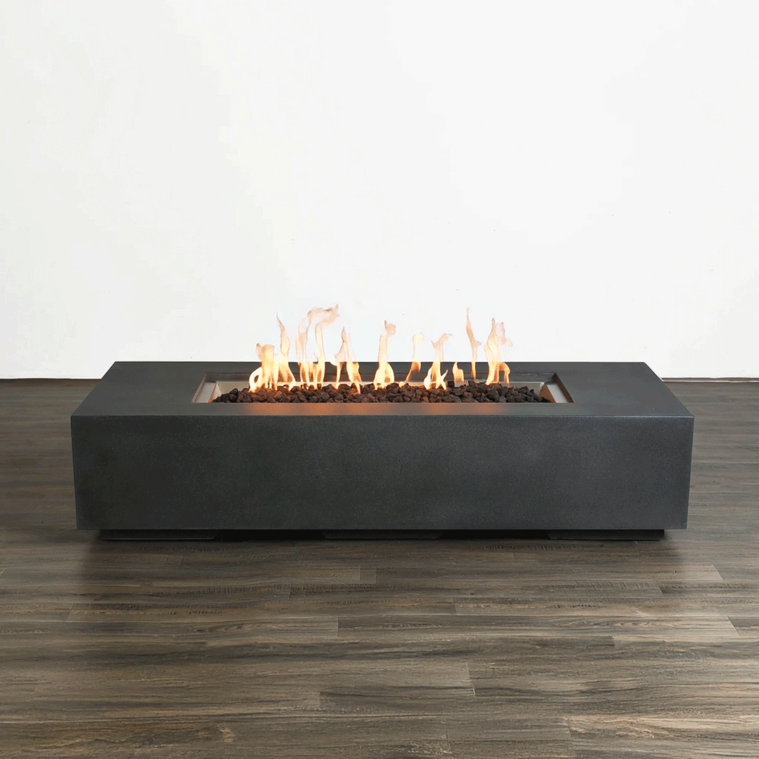 This studio view displays a modern fire pit with a sleek, graphite-colored finish resting on a dark wood floor. Bright orange flames rise continuously from a bed of black lava rocks, demonstrating the clean burn of a gas fire pit. The minimalist design of the linear fire pit is emphasized against a plain white background, showcasing the consistent flame pattern and architectural form without environmental distractions.