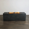 A studio presentation highlights a rectangular concrete fire pit with a matte graphite finish, resting on a dark wood floor against a white wall. Bright orange flames flicker consistently along the length of the linear fire pit, rising from a bed of black lava rocks. The minimalist setting emphasizes the clean lines and steady combustion of this natural gas fire pit, demonstrating its suitability for modern design aesthetics.