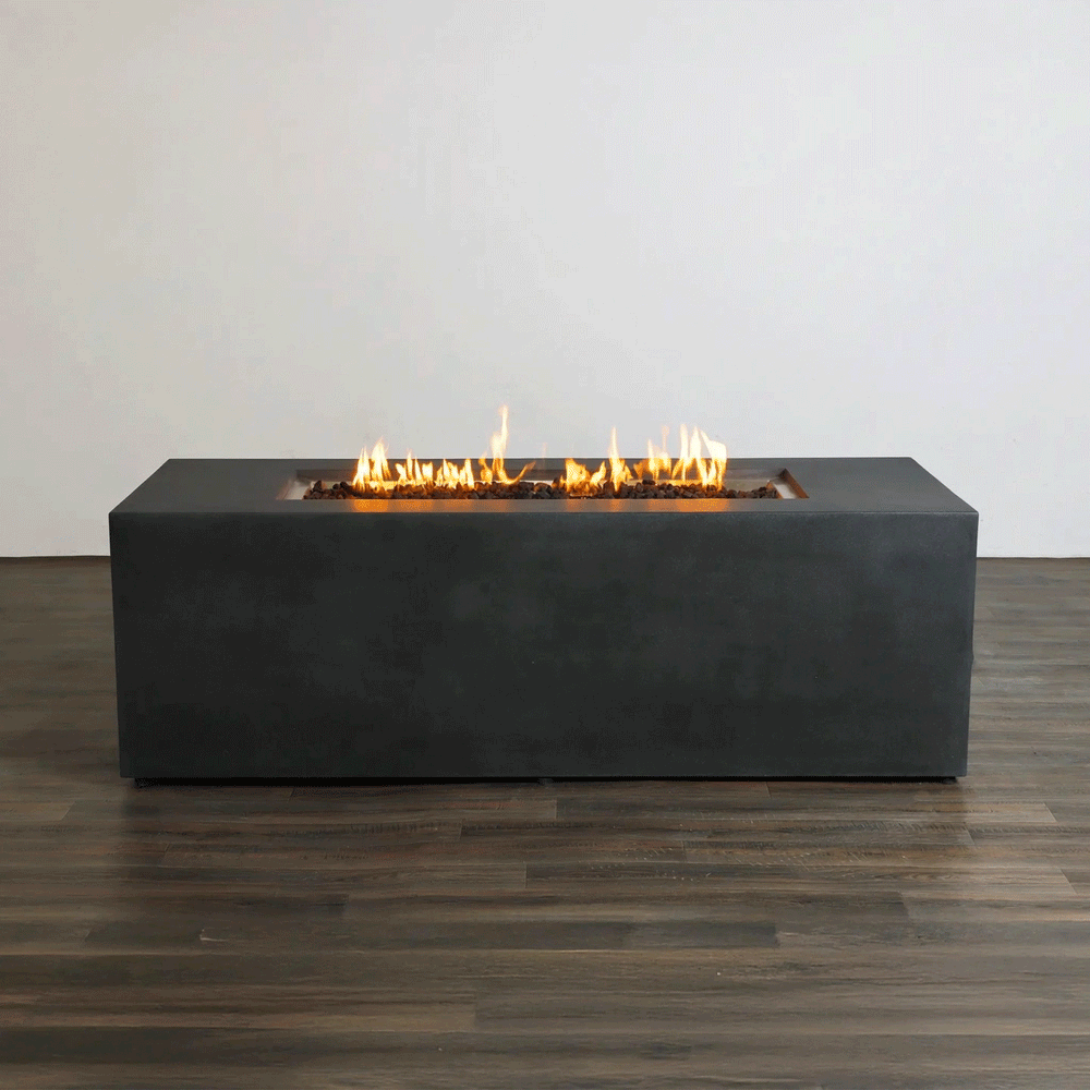 A studio presentation highlights a rectangular concrete fire pit with a matte graphite finish, resting on a dark wood floor against a white wall. Bright orange flames flicker consistently along the length of the linear fire pit, rising from a bed of black lava rocks. The minimalist setting emphasizes the clean lines and steady combustion of this natural gas fire pit, demonstrating its suitability for modern design aesthetics.