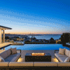 A luxury fire pit anchors a high-end terrace setting at twilight, positioned between large cushioned sofas and an infinity pool overlooking a bay and city skyline. The rectangular graphite fire table emits a warm glow that contrasts with the blue evening light, creating a sophisticated atmosphere for outdoor entertaining. Steady flames burn cleanly from the gas fire pit, enhancing the serene ambiance of this hillside estate.