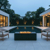 A symmetrical backyard fire pit area features a rectangular unit in a graphite finish, positioned before a swimming pool and modern pool house under a deep blue evening sky. Two upholstered armchairs face the central patio fire pit, where steady flames burn cleanly, illustrating the benefits of a smokeless fire pit. The scene highlights the architectural balance and warm lighting of a modern residential courtyard using a propane fire pit.