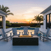 A rectangular fire pit with a graphite finish sits centrally on a paved pool deck, surrounded by four grey Adirondack chairs. The setting features palm trees and a vibrant sunset, creating an inviting space for a family gathering fire pit setup. Bright yellow flames rise from the fire feature, reflecting off the swimming pool water and demonstrating the unit's ability to anchor a tropical outdoor living space.