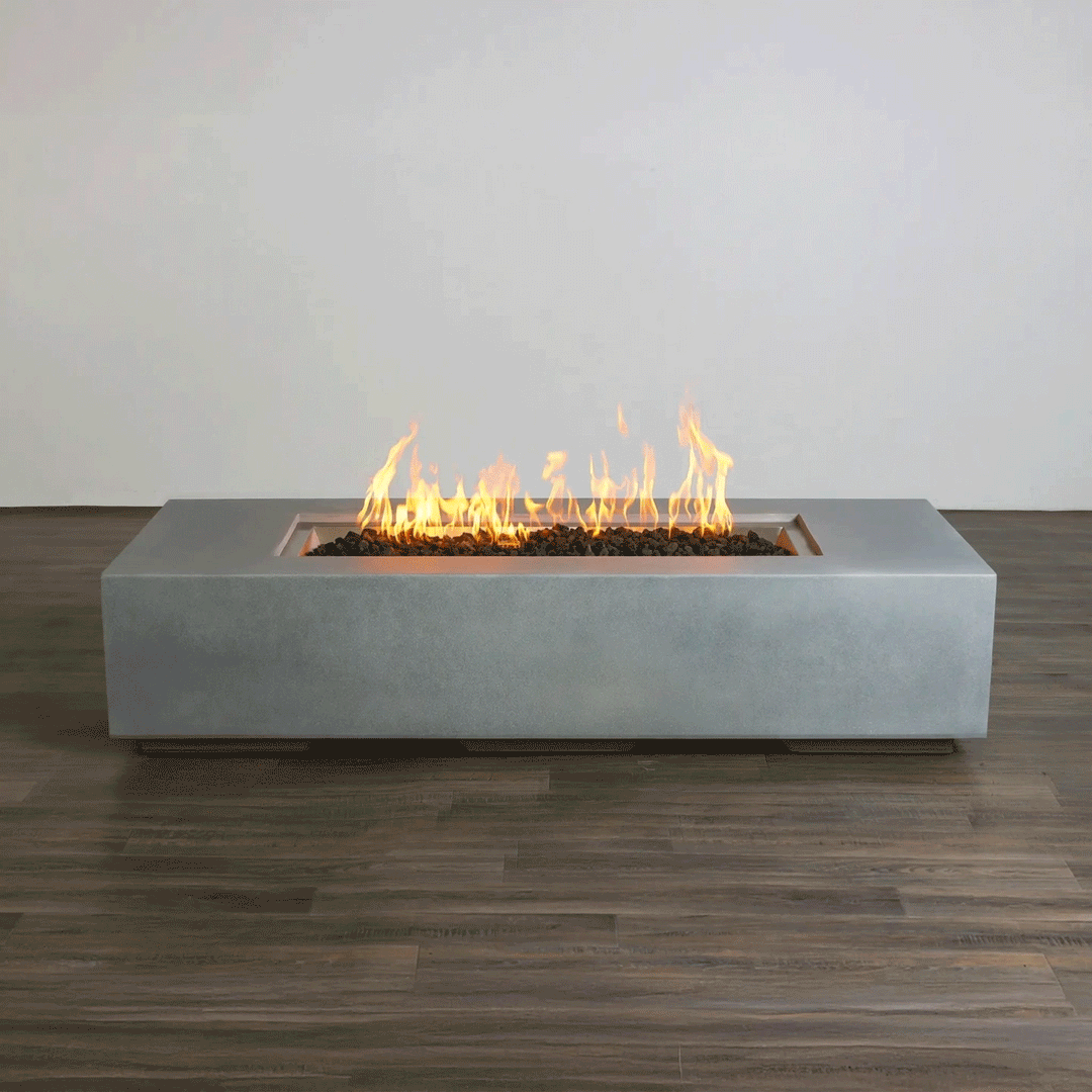 A studio presentation highlights a rectangular concrete fire pit with a smooth grey finish, resting on a dark wood floor against a white wall. Yellow and orange flames flicker consistently along the length of the linear fire pit, rising from a bed of dark lava media. The minimalist setting emphasizes the clean lines and steady combustion of this modern fire pit, demonstrating the aesthetic appeal of a propane fire pit solution for contemporary spaces.