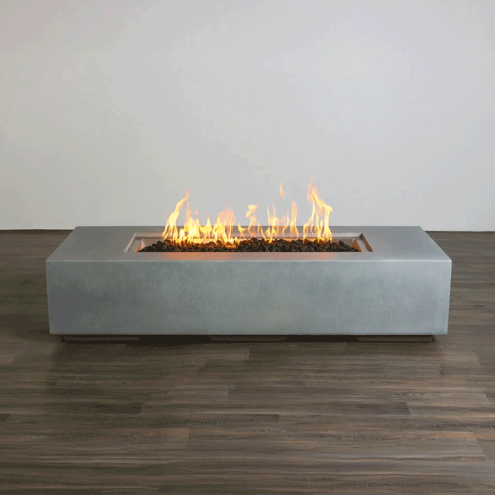A studio presentation highlights a rectangular concrete fire pit with a smooth grey finish, resting on a dark wood floor against a white wall. Yellow and orange flames flicker consistently along the length of the linear fire pit, rising from a bed of dark lava media. The minimalist setting emphasizes the clean lines and steady combustion of this modern fire pit, demonstrating the aesthetic appeal of a propane fire pit solution for contemporary spaces.