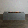 A studio presentation displays a rectangular concrete fire pit in a cool gray finish, resting on a dark wood floor against a plain white wall. Bright orange flames rise continuously from a bed of dark media along the length of the linear fire pit. The minimalist backdrop emphasizes the smooth texture and steady flame height, demonstrating the clean aesthetic of a natural gas fire pit connection suitable for a modern fire pit design.