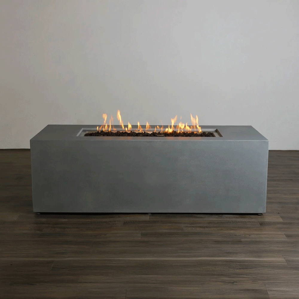 A studio presentation displays a rectangular concrete fire pit in a cool gray finish, resting on a dark wood floor against a plain white wall. Bright orange flames rise continuously from a bed of dark media along the length of the linear fire pit. The minimalist backdrop emphasizes the smooth texture and steady flame height, demonstrating the clean aesthetic of a natural gas fire pit connection suitable for a modern fire pit design.
