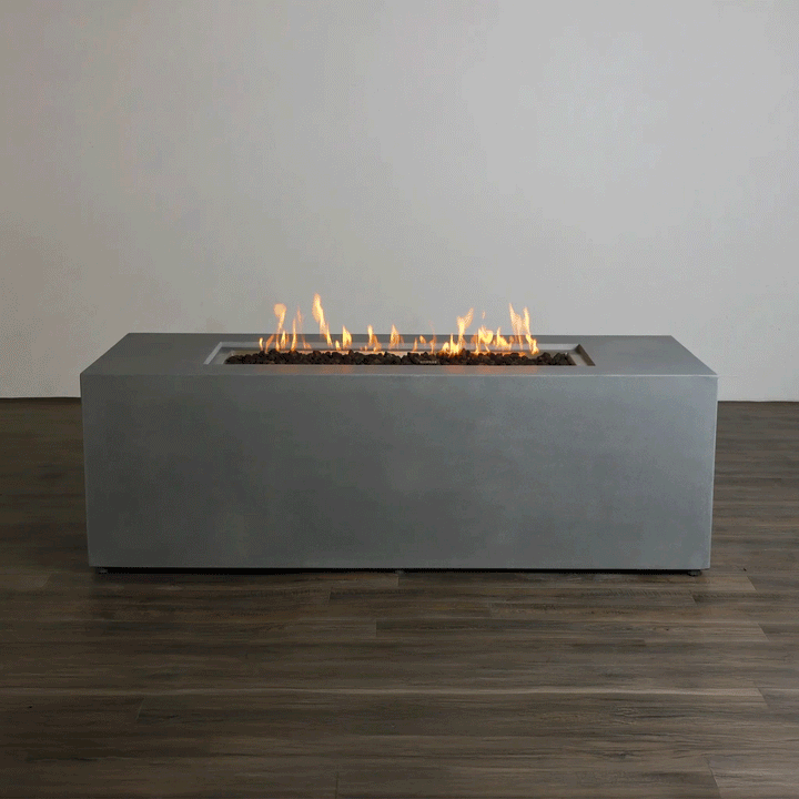 A studio presentation displays a rectangular concrete fire pit in a cool gray finish, resting on a dark wood floor against a plain white wall. Bright orange flames rise continuously from a bed of dark media along the length of the linear fire pit. The minimalist backdrop emphasizes the smooth texture and steady flame height, demonstrating the clean aesthetic of a natural gas fire pit connection suitable for a modern fire pit design.