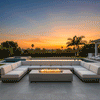 A spacious outdoor lounge features a large U-shaped sectional sofa surrounding a low-profile concrete fire pit in a grey finish. Set against a sunset backdrop with palm trees, the scene epitomizes a comfortable family gathering fire pit arrangement. The long, consistent flame pattern of the linear fire pit provides a warm focal point for the tiled patio, showcasing the sleek, geometric design of this modern fire pit setup.