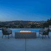 A rectangular outdoor fire pit in a gray finish sits on a concrete patio, flanked by two modern woven armchairs overlooking a rolling landscape at twilight. The fire table emits a warm glow against the deep blue sky, creating a cozy backyard fire pit atmosphere next to an infinity pool. The steady flames from the propane fire pit highlight the sleek design, offering a perfect spot for intimate conversation and relaxation.