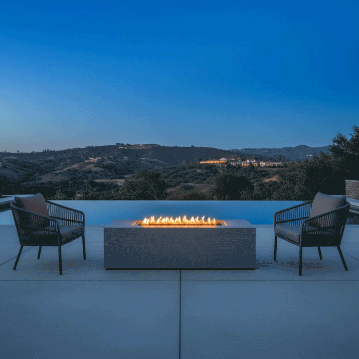 A rectangular outdoor fire pit in a gray finish sits on a concrete patio, flanked by two modern woven armchairs overlooking a rolling landscape at twilight. The fire table emits a warm glow against the deep blue sky, creating a cozy backyard fire pit atmosphere next to an infinity pool. The steady flames from the propane fire pit highlight the sleek design, offering a perfect spot for intimate conversation and relaxation.
