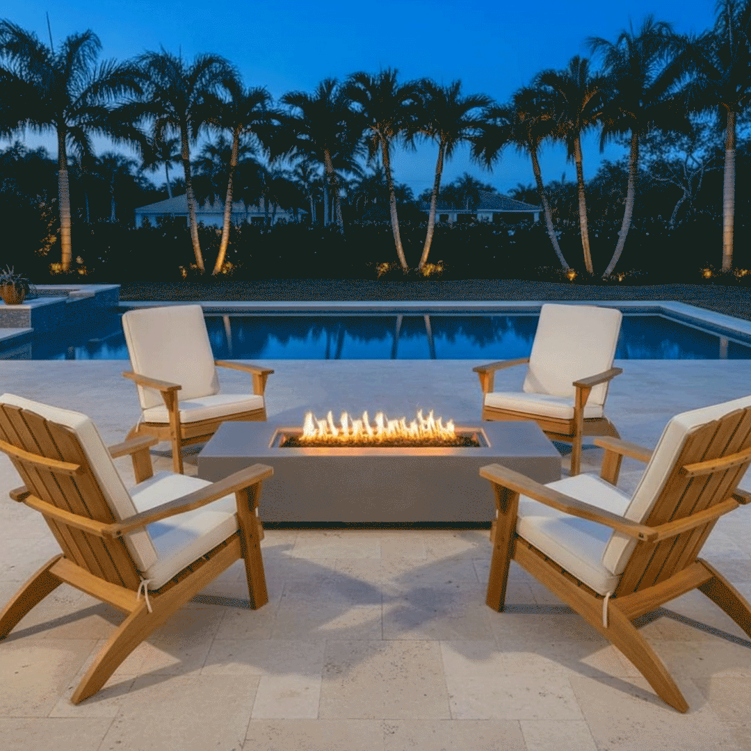 A twilight scene displays a grey patio fire pit positioned centrally on a stone deck, surrounded by tall palm trees and a swimming pool. Four wooden armchairs with white cushions face the rectangular unit, creating a perfect symmetrical layout for outdoor entertaining. The steady flames illuminate the backyard oasis, reflecting softly against the water and enhancing the tropical ambiance of the outdoor fire pit area.