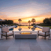 A luxury fire pit anchors a serene terrace setting during a golden sunset, flanked by two cushioned armchairs and an infinity pool overlooking the ocean. The rectangular grey structure houses a vibrant fire feature that burns cleanly without visible smoke, demonstrating the efficiency of a smokeless fire pit. The warm glow of the flames complements the orange sky, creating an inviting, high-end atmosphere around this gas fire pit.