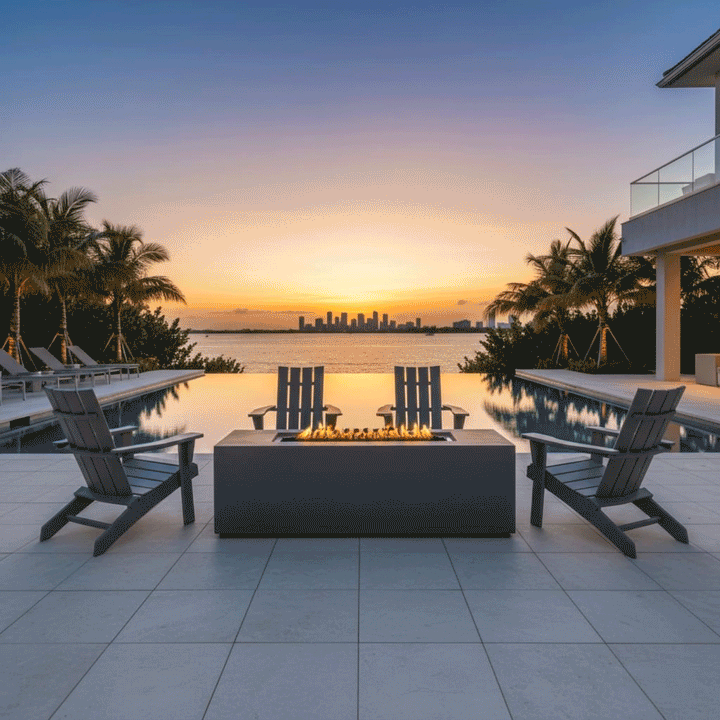 A gray patio fire pit serves as the centerpiece of a waterfront terrace at sunset, surrounded by four gray Adirondack chairs. The fire feature burns cleanly with golden flames, demonstrating the benefits of a smokeless fire pit in a breezy coastal setting. Positioned near an infinity pool with palm trees, this gas fire pit creates a serene ambiance, reflecting the warm colors of the sky and enhancing the luxury of the outdoor deck.