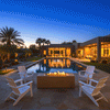 A propane fire pit in a warm sand tone sits on a flagstone patio surrounded by four white Adirondack chairs. The scene features a large swimming pool and a modern desert-style home illuminated at twilight. Steady flames from the fire feature create an inviting atmosphere for a family gathering fire pit setup, highlighting the geometric design of this backyard fire pit area.