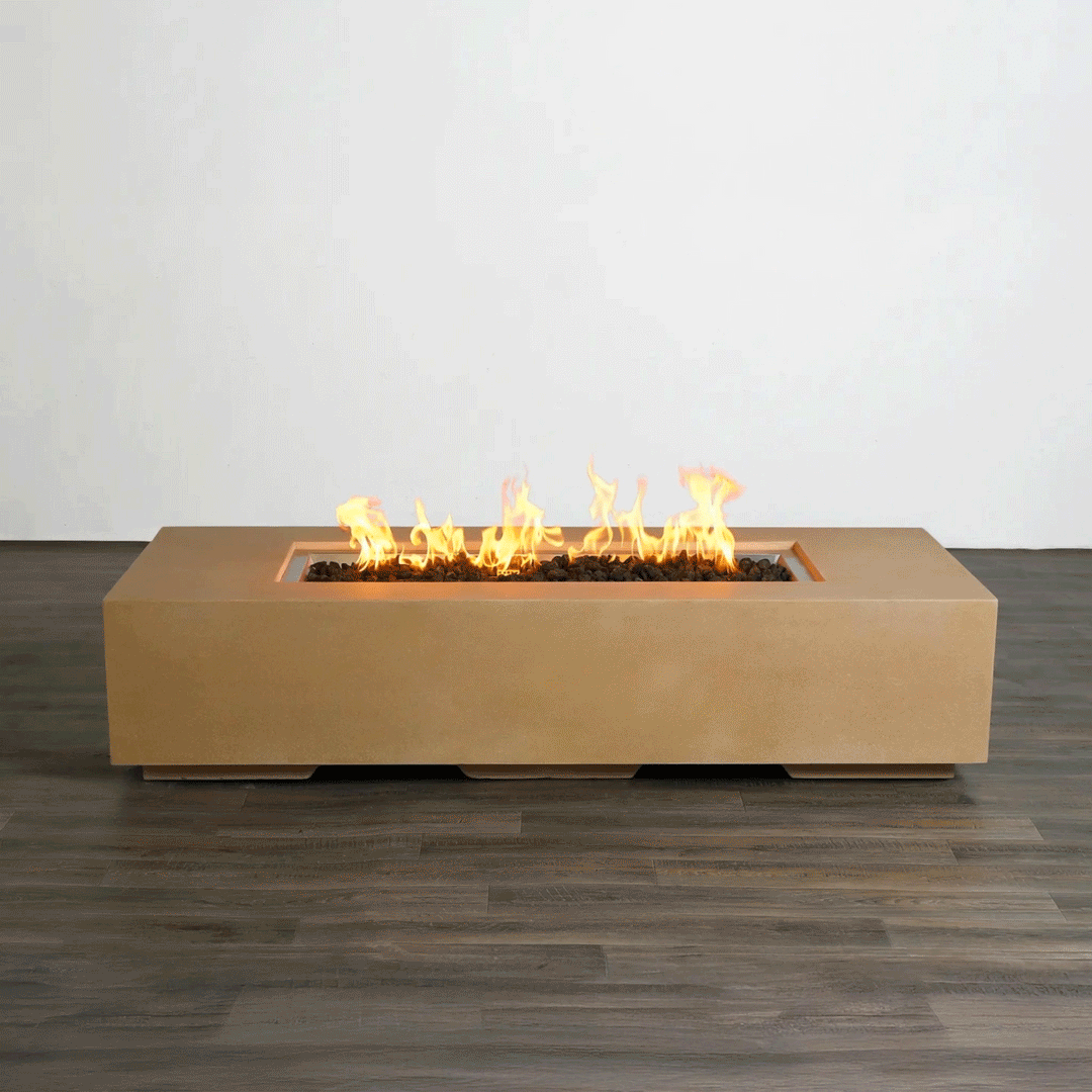 A studio view presents a rectangular concrete fire pit in a natural sand finish, contrasting against a dark wood floor and white background. Bright orange flames rise continuously from a bed of black lava rocks along the length of the linear fire pit, demonstrating the clean, smoke-free combustion. The minimalist presentation emphasizes the smooth texture and steady flame height typical of a high-quality natural gas fire pit connection.