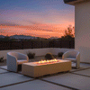 A modern fire pit in a warm beige tone sits centrally on a paved terrace, blending with the desert landscape and distant mountains during a vibrant sunset. Two curved armchairs face the rectangular fire pit, creating an intimate conversation spot. The flames burn cleanly without obstructing the scenic view, showcasing the benefits of a smokeless fire pit as a central patio fire pit feature in an arid, open-air setting.