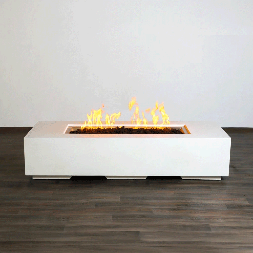 A low white concrete fire pit with crisp edges stretches horizontally across a dark wood-look floor indoors, its long recessed channel packed with rough lava rocks while bright yellow flames from the gas fire pit rise and fall in a slow rhythm, clearly showing the elongated rectangular fire pit form and raised plinth base that give this minimalist modern fire pit a clean, sculptural presence against the plain light wall.