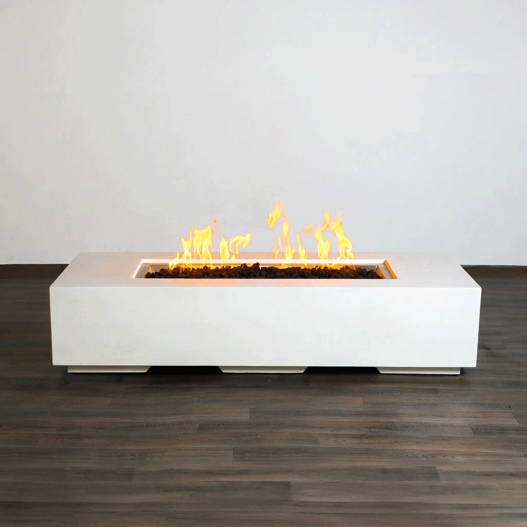 A low white concrete fire pit with crisp edges stretches horizontally across a dark wood-look floor indoors, its long recessed channel packed with rough lava rocks while bright yellow flames from the gas fire pit rise and fall in a slow rhythm, clearly showing the elongated rectangular fire pit form and raised plinth base that give this minimalist modern fire pit a clean, sculptural presence against the plain light wall.