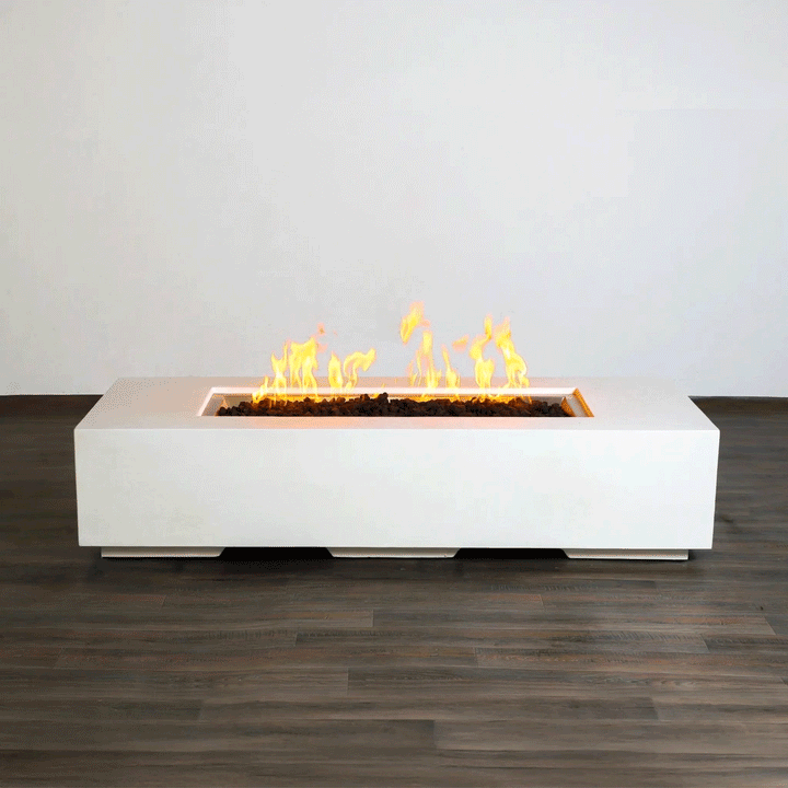 A low white concrete fire pit with crisp edges stretches horizontally across a dark wood-look floor indoors, its long recessed channel packed with rough lava rocks while bright yellow flames from the gas fire pit rise and fall in a slow rhythm, clearly showing the elongated rectangular fire pit form and raised plinth base that give this minimalist modern fire pit a clean, sculptural presence against the plain light wall.