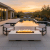 On a pale stone terrace beside a still swimming pool reflecting a warm orange sunset, a white concrete fire pit with a narrow rectangular opening burns in a steady line of flames between two cushioned lounge chairs, while desert plants, barrel cacti, and distant mountains frame the horizon, creating an inviting outdoor fire pit scene that reads as a calm backyard oasis designed for quiet evening conversation and poolside relaxation.