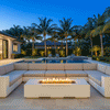 A long white concrete fire pit sits at the center of a U-shaped sectional sofa on a tiled patio, its continuous row of flames flickering above dark lava rocks as palm trees and landscaped greenery around the nearby pool glow with soft evening uplighting, depicting a modern fire pit arrangement that naturally becomes the focus of the outdoor fire pit seating zone, suited to relaxed outdoor entertaining and group gatherings.