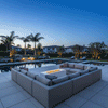 A large U-shaped sectional sofa surrounds a low-profile white fire table, creating a spacious family gathering fire pit area. Set within a backyard oasis featuring tall palm trees and a reflective swimming pool, the scene captures a serene twilight ambiance. The steady, clean-burning flames of the propane fire pit provide warmth and light, anchoring the extensive outdoor lounge space.