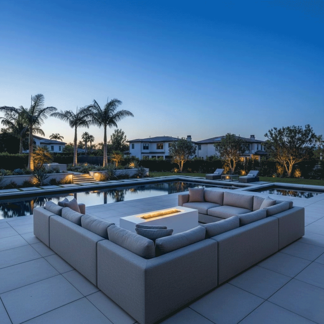 A large U-shaped sectional sofa surrounds a low-profile white fire table, creating a spacious family gathering fire pit area. Set within a backyard oasis featuring tall palm trees and a reflective swimming pool, the scene captures a serene twilight ambiance. The steady, clean-burning flames of the propane fire pit provide warmth and light, anchoring the extensive outdoor lounge space.