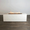 A studio view presents a rectangular concrete fire pit in a stark white finish, contrasting with a dark wood floor. Bright orange flames rise continuously from a bed of black lava rocks along the length of the linear fire pit. The minimalist backdrop emphasizes the architectural form and the consistent flame height typical of a premium natural gas fire pit connection.