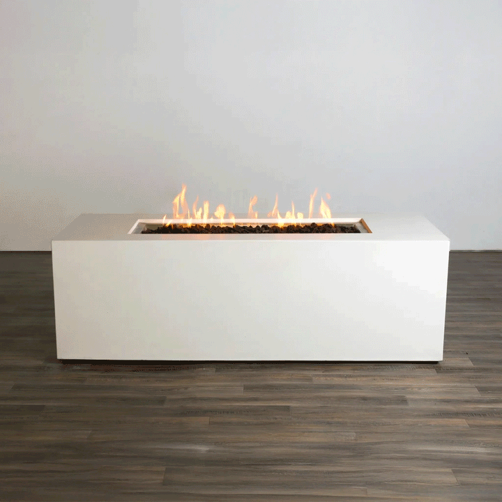 A studio view presents a rectangular concrete fire pit in a stark white finish, contrasting with a dark wood floor. Bright orange flames rise continuously from a bed of black lava rocks along the length of the linear fire pit. The minimalist backdrop emphasizes the architectural form and the consistent flame height typical of a premium natural gas fire pit connection.
