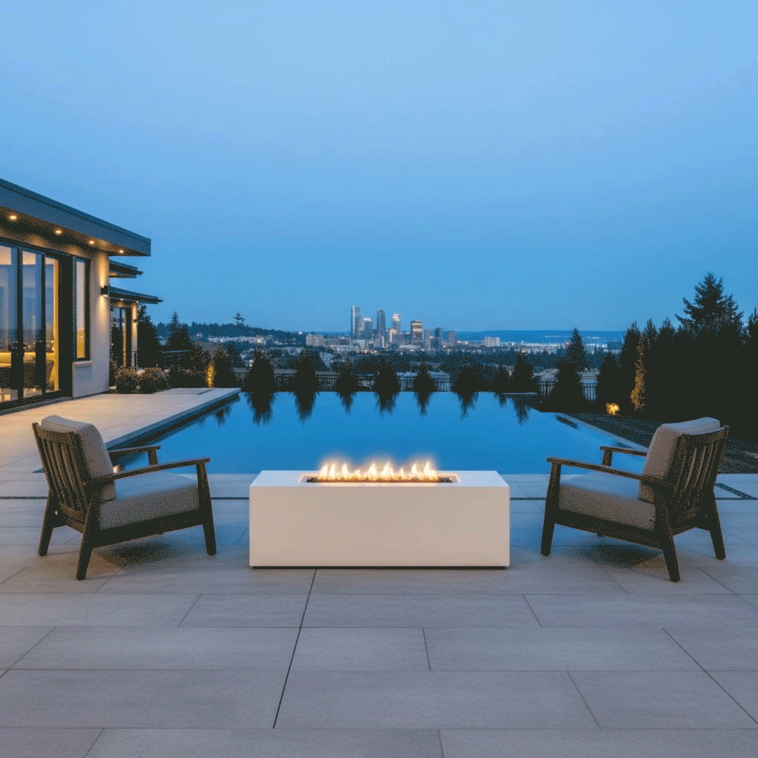 A luxury fire pit in a white finish sits on a tiled terrace edge, flanked by two armchairs overlooking an infinity pool and a distant city skyline. The blue evening light contrasts with the warm amber flames of the gas fire pit, which burn cleanly to demonstrate a smokeless fire pit experience. The setting highlights a sophisticated spot for outdoor entertaining with a panoramic backdrop.