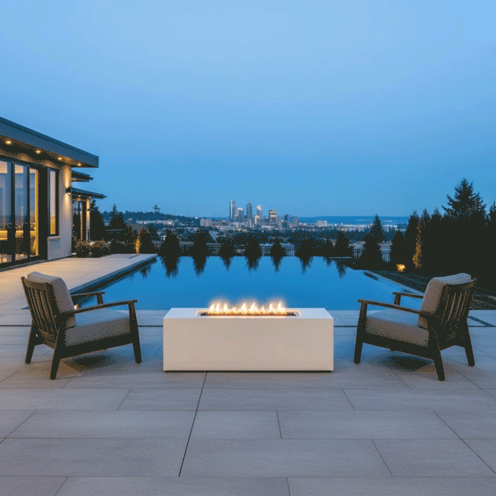 A luxury fire pit in a white finish sits on a tiled terrace edge, flanked by two armchairs overlooking an infinity pool and a distant city skyline. The blue evening light contrasts with the warm amber flames of the gas fire pit, which burn cleanly to demonstrate a smokeless fire pit experience. The setting highlights a sophisticated spot for outdoor entertaining with a panoramic backdrop.