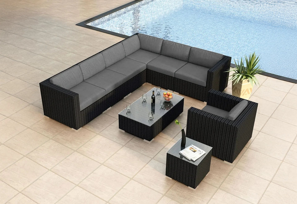 Modern black wicker outdoor sectional set with gray cushions, featuring a glass-top coffee table and accent table, arranged on a poolside patio with a stylish planter.