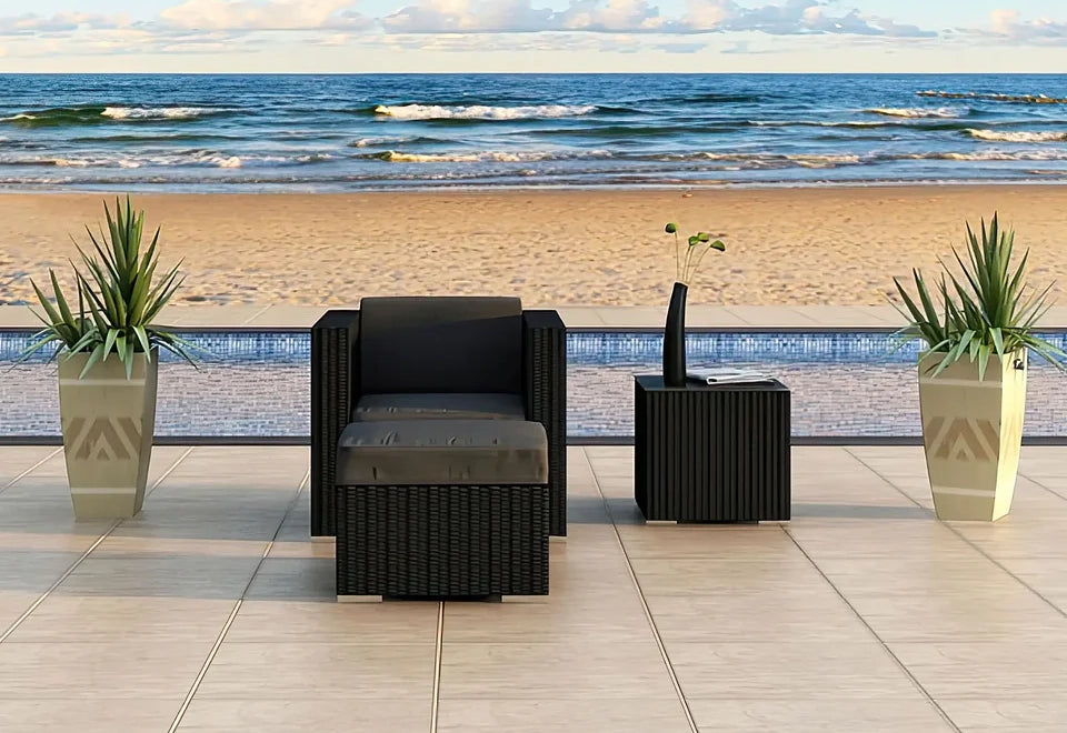 Black wicker patio chair with dark gray cushions paired with a matching square coffee table, set against a serene beachfront backdrop with potted greenery