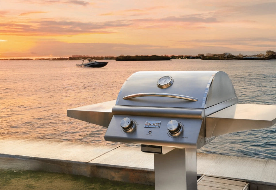 Blaze stainless steel post-mount grill with dual burners and lid, set against a serene waterfront sunset backdrop, ideal for outdoor cooking.