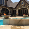 A luxurious estate-style backyard with two Cazo 24" Round Concrete Fire Bowls by The Outdoor Plus, positioned above a light-colored pool. The bowls, in a warm sand-toned finish, display glowing flames and rest on elegant curved pedestals, perfectly complementing the home’s stone facade and grand architectural design.
