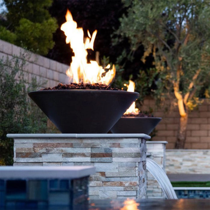 A dramatic evening setting featuring The Outdoor Plus Cazo 24" Round Fire Bowl in a deep chocolate finish. Flames rise from lava rock as water flows from the built-in scupper into a serene pool below. The stone pedestal and surrounding landscaping create a peaceful and upscale outdoor retreat.