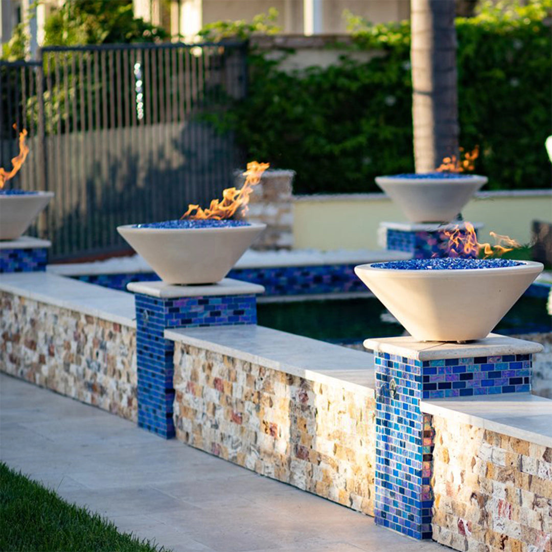 A stylish row of The Outdoor Plus Cazo 24" Round Concrete Fire Bowls in a soft Vanilla finish, each filled with blue fire glass and vibrant flames. Mounted on mosaic-tiled pillars along a backyard pool, these bowls create a stunning visual rhythm and elevate the ambiance of the outdoor space with warmth and elegance.