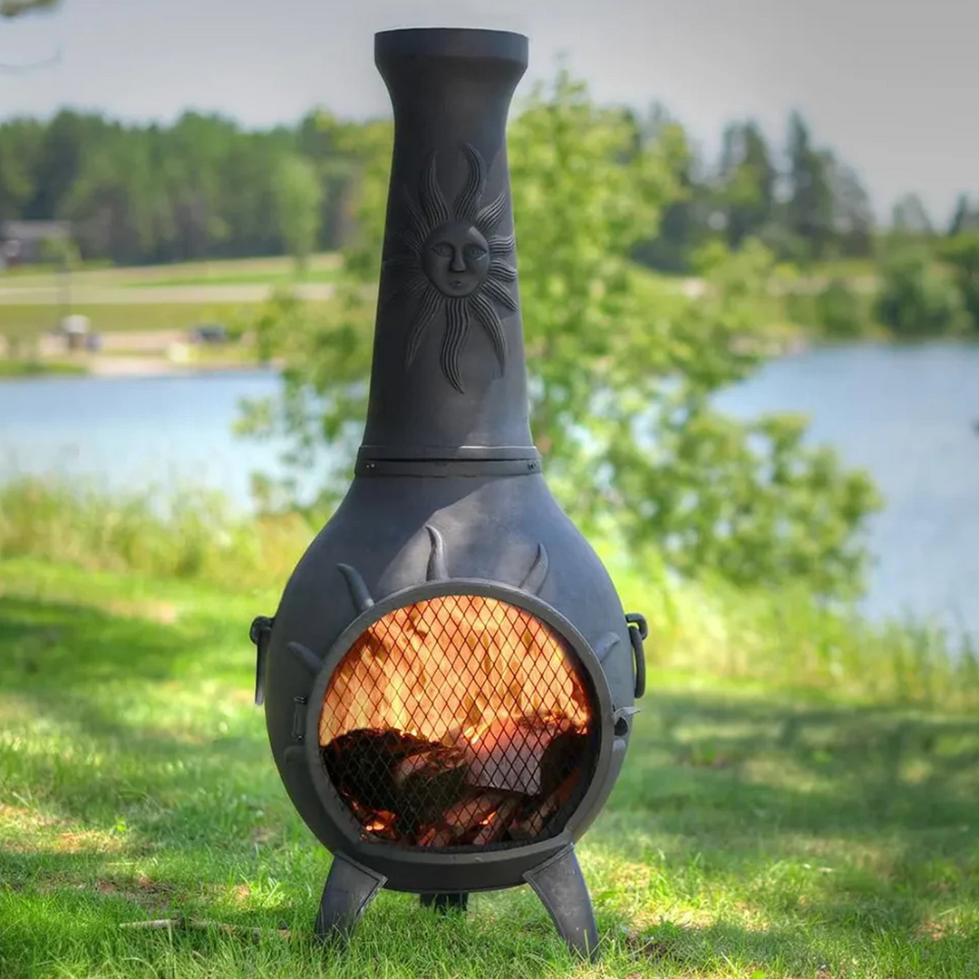 Black cast iron chiminea with a sun design on the chimney, burning wood inside, placed on a grassy area near a lake with trees in the background