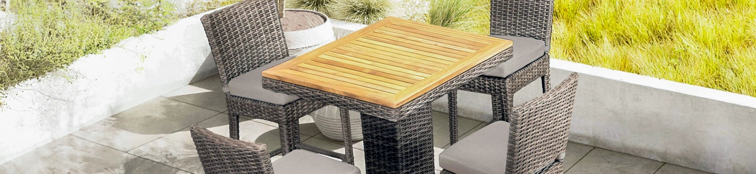 Stylish outdoor bar-height bistro set featuring a wooden slatted table and wicker chairs with cushioned seats, arranged on a modern patio with a backdrop of greenery, perfect for casual outdoor dining or entertaining